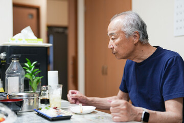In a small Japanese apartment, a Japanese man in his late 70s relaxes in T-shirt pajamas. He sits alone in the evening, sipping whiskey and enjoying a calm, quiet moment after a long day of solitude.