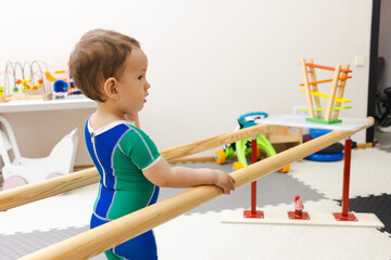 Little male child with movement disorders while participating in physical rehabilitation standing near rehab handrails in vibrant recovery space