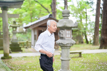 Fototapeta premium In rural Japan, a Japanese man in his late 70s visits a countryside shrine surrounded by lush green trees. Wearing a white shirt and hat, he prays quietly in the peaceful warmth of early summer air.