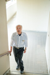 In Japan, a Japanese man in his late 70s, wearing a white shirt, climbs the stairs of an apartment using the handrail for support. This peaceful moment highlights aging with dignity and the quiet stre