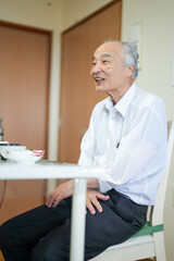 In Japan, a Japanese man in his late 70s wearing a white shirt is sitting and talking in a modest...