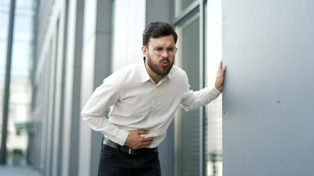Businessman feels stomach pain while standing on street near business office building. Sick man has heartburn, gastritis, bloating or poisoning. An unhealthy male suffers from spasms and constipation