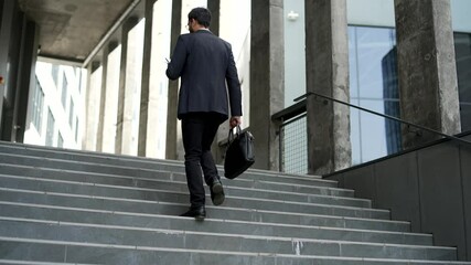 Businessman walking up stairs of office building holding smartphone. Confident entrepreneur in formal suit with briefcase going to work checking phone in urban city. Professional business. Rear view - Powered by Adobe