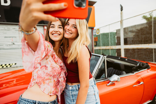 Two young women taking selfie with red convertible car