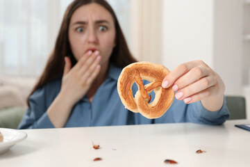 Shocked young woman holding pretzel with cockroach at white table indoors, selective focus. Pest...