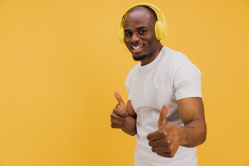 Young man listening music with yellow headphones showing thumbs up on yellow background