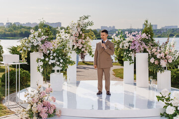 A man presenter in a beige suit leads a wedding ceremony in the park. 