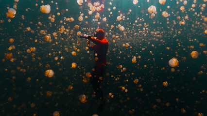 Male freediver in red wetsuit swims underwater in the lake full of jellyfish. West Papua, Raja Ampat, Indonesia