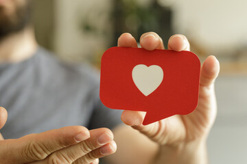 Close-up of a man's hands gently holding a red speech bubble with a white heart, symbolizing social...