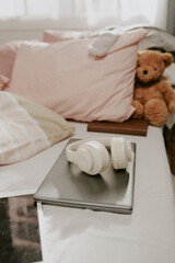 Headphones resting on laptop on bed with plush bear, creating a cozy scene. Sunlight filtering through a window, illuminating the soft bedding and creating a warm ambiance
