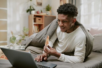 African American man enjoying slice of pizza while sitting in bed and smiling at laptop. Cozy...