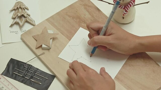Overhead view of drawing and marking a star shape on a white paper to design a Christmas card surrounded by festive items and decorations to show holidays crafts