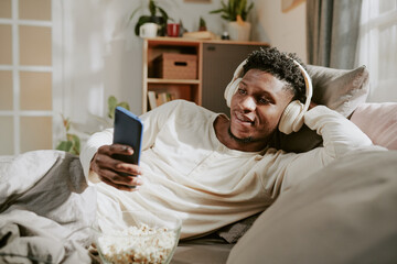 African American man lying on couch wearing headphones and holding a smartphone while leaning back....