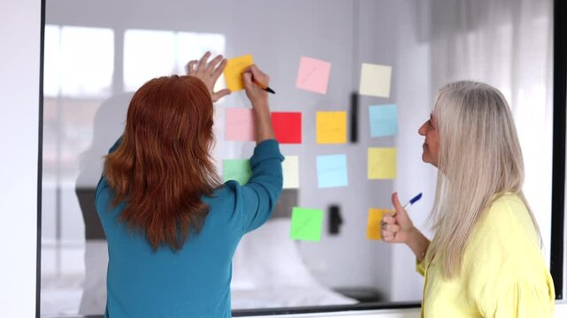 Professional businesswomen strategizing and brainstorming, using colorful sticky notes on glass wall for innovative project planning and collaborative problem solving in modern office environment