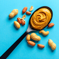 A spoon with smooth peanut butter surrounded by whole and shelled peanuts on a bright blue background