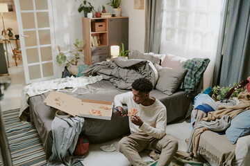 Multiracial man sitting on floor, painting in sketchbook in a comfortably cluttered living room...