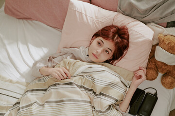Young girl with red hair lying in bed under a striped blanket while holding it with a thoughtful...