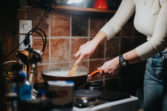 A woman stirring food in a pan on a stove top with wooden cabinets and tiles in the background, showcasing a kitchen environment for preparing meals.