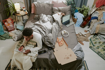 Person lying on bed and using tablet while eating pizza with various items scattered around,...