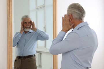 Handsome senior man looking at himself in mirror indoors