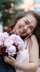 Beautiful woman with bouquet of peonies talking on phone and looking happy outdoors. Urban...