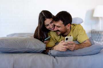 Happy couple lying on the bed, smiling and using smartphones, enjoying moments of connection and...