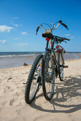 Fototapeta premium Two bicycles parked on a sandy beach with the sea and clear blue sky. Cycling to the beach on a bike for vacation leisure activity by the water. Swimming, sunbathing and relaxing