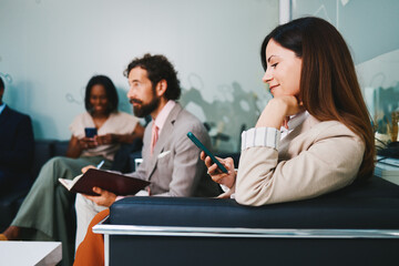 Businesswoman smiling and using smartphone while waiting in office lobby with colleagues