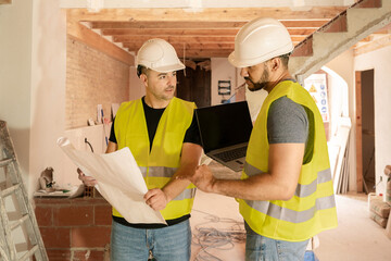 Two Construction workers analyzing building plans with laptop on construction site