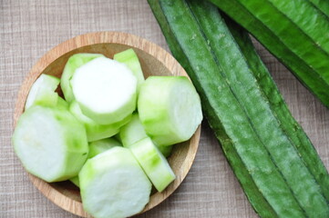 Whole, green ridge gourds (Luffa acutangula) next to a small wooden bowl filled with several slices...