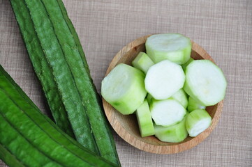 Whole, green ridge gourds (Luffa acutangula) next to a small wooden bowl filled with several slices...