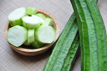 Whole, green ridge gourds (Luffa acutangula) next to a small wooden bowl filled with several slices...