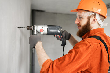 Portrait of concentrated bearded construction worker drilling hole with electric drill in wall....