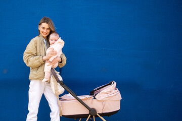 Smiling young mother holding baby by a pink stroller against a blue wall.