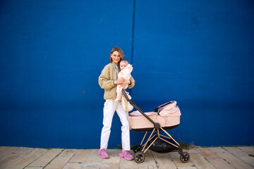 Full body young mother holding her baby beside a pink stroller, standing against blue wall
