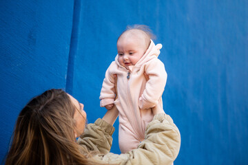 Mother lifting and playing with her smiling baby dressed in a soft pink outfit, set against a vibrant blue wall.