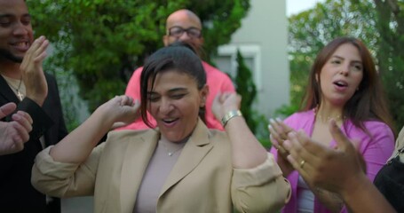 Woman raising fists in celebration while surrounded by friends clapping in outdoor gathering, joyful expression as she reacts to recognition and shared excitement