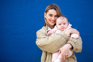Smiling mother holding her baby in a pink fleece outfit against a bright blue wall