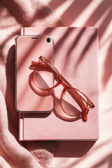 Smartphone and eyeglasses on pink notebook with soft shadows. Modern workspace with minimal accessories on desk