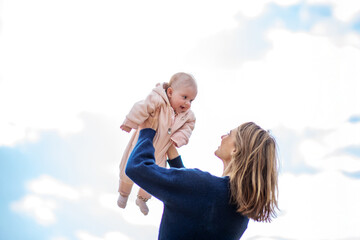 Cheerful mother lifts her baby into the air against a bright sky