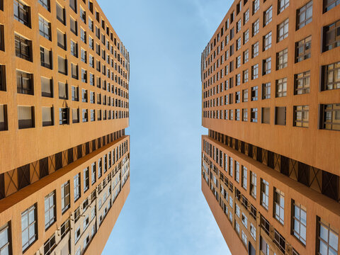 Striking upward view of two symmetrical modern high-rise buildings with orange facades, showcasing urban architecture, geometry, and city living under a clear blue sky.