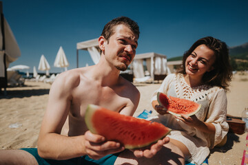 A couple sharing watermelon slices while relaxing on a sandy beach under the bright sun. The atmosphere conveys happiness, relaxation, and the essence of summer recreation by the seaside.