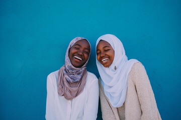 Two happy african american teenagers laughing together. Smiling black muslim female friends. Blue wall. Candid authentic inclusive friendship. Happy ramadan