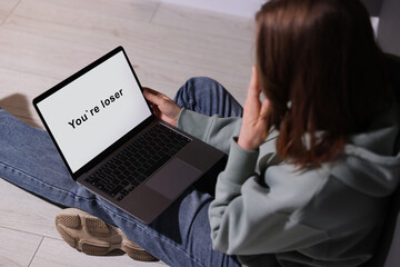Cyber bullying. Upset teenage girl looking at laptop with text You Loser on floor indoors, closeup