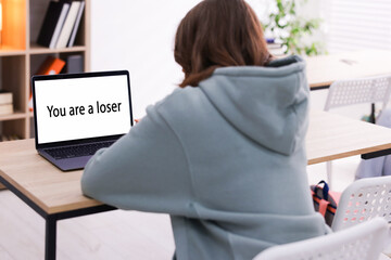 Cyber bullying. Upset teenage girl and laptop with text You Loser at desk indoors