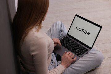 Cyber bullying. Upset teenage girl looking at laptop with text You Loser on floor indoors, closeup