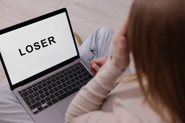 Cyber bullying. Upset teenage girl looking at laptop with text You Loser on floor indoors, closeup