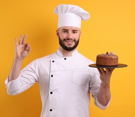 Happy confectioner in uniform holding chocolate cake and showing ok gesture on orange background