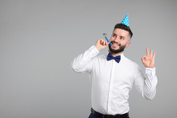 Happy Birthday. Handsome man with party hat and blower showing ok gesture on grey background, space for text