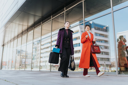Two women in professional attire walking together near an office building, discussing work and holding accessories such as bags and documents, symbolizing collaboration and modern business.